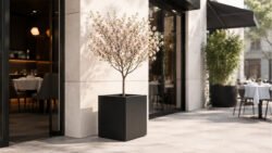 Schwarzer, quadratischer Aluminium-Pflanzkübel Alessio 1 mit kleinem blühendem Baum vor dem hellen Eingang eines modernen Restaurants