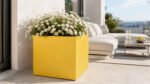 Gelber, quadratischer Pflanzkübel Alessio 5 aus Aluminium mit weißen Margeriten neben einer hellen Lounge-Ecke auf einer sonnigen Terrasse