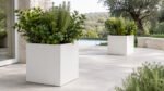 Zwei weiße Pflanzkübel Alessio 6 aus Aluminium mit üppiger grüner Bepflanzung auf einer hellen Gartenterrasse, ein Kübel im Vordergrund und ein weiterer im Hintergrund