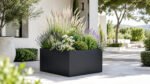 Schwarzer, großformatiger Pflanzkübel Alessio 8 aus Aluminium mit Ziergräsern, Lavendel, weißen Blüten und kompakten Grünpflanzen auf einer hellen Gartenterrasse