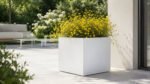 Großer, quadratischer Pflanzkübel Alessio 10 aus weißem Aluminium mit zarten gelben Blüten auf einer hellen, modernen Gartenterrasse