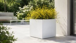 Großer, quadratischer Pflanzkübel Alessio 10 aus weißem Aluminium mit zarten gelben Blüten auf einer hellen, modernen Gartenterrasse