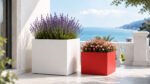 Weißer Pflanzkübel Alessio 10 aus Aluminium mit Lavendel neben einem roten Pflanzkübel mit farbenfrohen Blüten auf einer hellen Terrasse mit Meerblick
