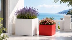 Weißer Pflanzkübel Alessio 10 aus Aluminium mit Lavendel neben einem roten Pflanzkübel mit farbenfrohen Blüten auf einer hellen Terrasse mit Meerblick