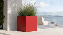 Roter, quadratischer Pflanzkübel Alessio 15 aus Aluminium mit üppigem Ziergras auf einer hellen Terrasse mit Glasgeländer und weitem Blick über das Wasser