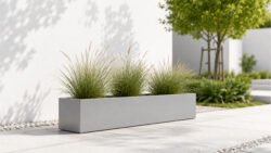 Hellgrauer, rechteckiger Pflanzkübel Alessio 9 aus Aluminium mit dekorativen Ziergräsern auf einem hellen Gartenweg in minimalistischer Umgebung