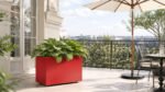 Roter Pflanzkübel Apollo 14 Mobile aus Aluminium mit grünen Hostas auf einem hellen Balkon neben einem Sonnenschirm und einer dekorativen Metallbalustrade