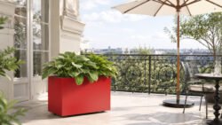 Roter Pflanzkübel Apollo 14 Mobile aus Aluminium mit grünen Hostas auf einem hellen Balkon neben einem Sonnenschirm und einer dekorativen Metallbalustrade