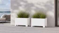 Zwei weiße Pflanzkübel aus Aluminium auf einer hellen Terrasse, nebeneinander platziert und mit dichter grüner Ziergras-Bepflanzung, vor ruhigem, modernem Hintergrund