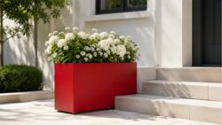 Roter Pflanzkübel aus Aluminium mit Rollen, bepflanzt mit weißen Blüten, dekorativ neben hellen Eingangsstufen an einer Hausfassade platziert