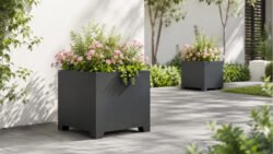 Zwei schwarze Apollo 19 Pflanzkübel aus Aluminium mit grünen Pflanzen und rosafarbenen Blüten auf hellem Gartenpflaster