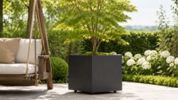 Schwarzer Apollo 19 Mobile Pflanzkübel aus Aluminium mit kleinem Zierbaum, auf heller Pflasterfläche im Garten neben einer Holzschaukel platziert