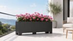 Schwarzer Apollo 20 Pflanzkübel aus Aluminium mit pinkfarbenen Blüten auf einem hellen Balkon mit Glasgeländer und weitem Ausblick