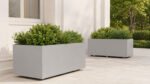 Zwei hellgraue Apollo 20 Mobile Pflanzkübel aus Aluminium mit grünen Sträuchern und Ziergräsern auf einer hellen Terrasse, einer im Vordergrund und einer weiter hinten platziert