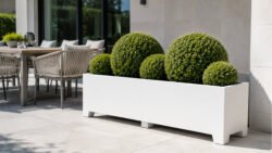Weißer Pflanzkübel Apollo 26 aus Aluminium mit kugelförmigen Buchsbaumsträuchern auf einer hellen Terrasse neben Tisch und Stühlen