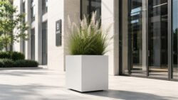 Weißer Pflanzkübel Apollo 4 Mobile aus Aluminium mit blühendem Ziergras vor einem hellen Bürogebäude