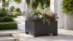 Apollo 5 Pflanzkübel aus schwarzem Aluminium auf kurzen Füßen, bepflanzt mit weißen, violetten und rosafarbenen Blüten, in einem hellen, modern gestalteten Garten