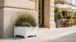 Weißer Pflanzkübel Apollo 5 aus Aluminium mit blühendem Ziergras und farblich abgestimmten Blumen neben einer Restaurantfassade