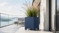 Apollo 7 Pflanzkübel aus blauem Aluminium auf Füßen, bepflanzt mit blühendem Ziergras, auf einem hellen Balkon mit Glasgeländer und Wasserblick platziert