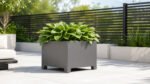 Grauer Pflanzkübel Apollo 8 Mobile aus Aluminium mit üppigen grünen Hostas auf einer hellen Terrasse vor einem dunklen Gartenzaun