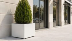 Weißer Pflanzkübel Apollo 8 Mobile aus Aluminium mit einer dichten grünen Thuja seitlich vor einem hellen Geschäftseingang