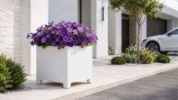 Apollo 9 Pflanzkübel aus weißem Aluminium auf Füßen, bepflanzt mit violetten Blüten, neben der Auffahrt vor einem hellen, modern gestalteten Haus platziert