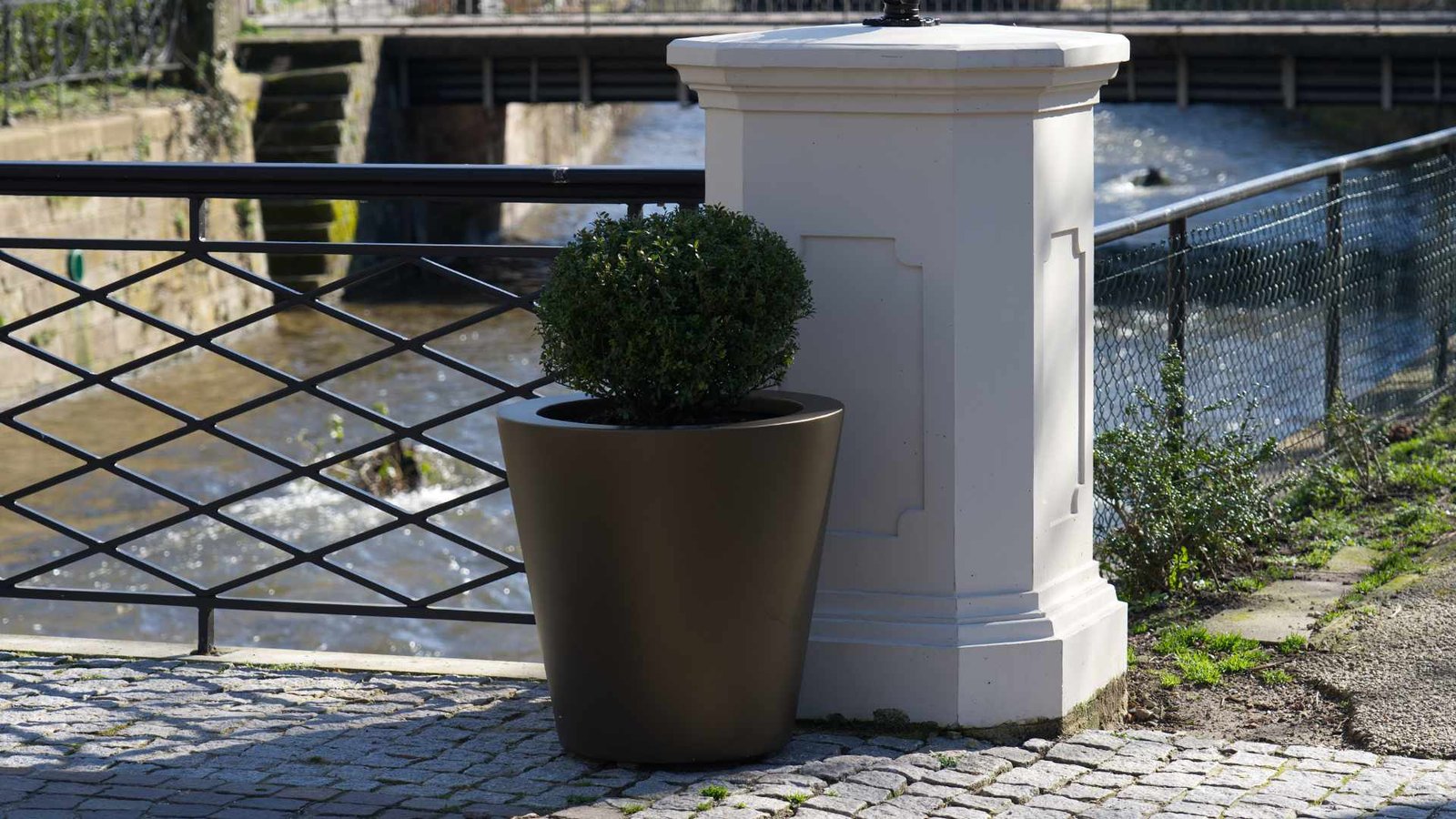 Pflanzkübel Aluminium in Bronze mit rund geschnittenem Buchsbaum auf einer gepflasterten Terrasse am Wasser.